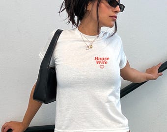 Puede incluir: Camiseta blanca con las palabras "House Wife" en letras rojas y un corazón rojo. La camiseta es de manga corta y parece ser de algodón. Se ve un bolso negro.