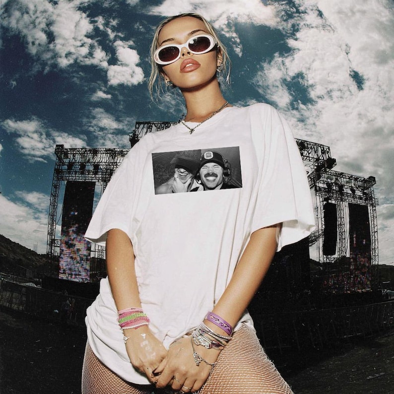 Może przedstawiać: Biały t-shirt z czarno-białym zdjęciem dw&oacute;ch os&oacute;b. Osoba nosi białe okulary przeciwsłoneczne i kilka bransoletek. Tło przedstawia zachmurzone niebo i scenę koncertową.