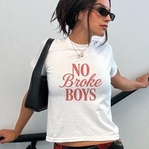 May include: White t-shirt with the phrase "NO Broke BOYS" in red script. The shirt is short-sleeved and has a crew neck. The model is wearing sunglasses and a black shoulder bag.