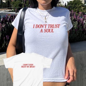 Puede incluir: Camiseta blanca con el texto rojo "I DON'T TRUST A SOUL". También se ve una segunda camiseta blanca con el texto "I DON'T EVEN TRUST MY BITCH!". Las camisetas son de manga corta.