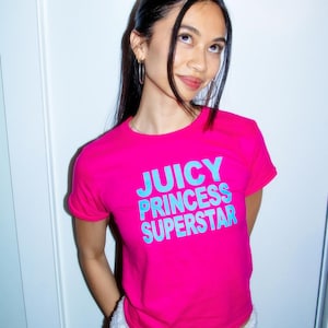 May include: A bright pink t-shirt with the text "JUICY PRINCESS SUPERSTAR" in light blue. The model is wearing white shorts. The image is well-lit and the colors are vibrant.