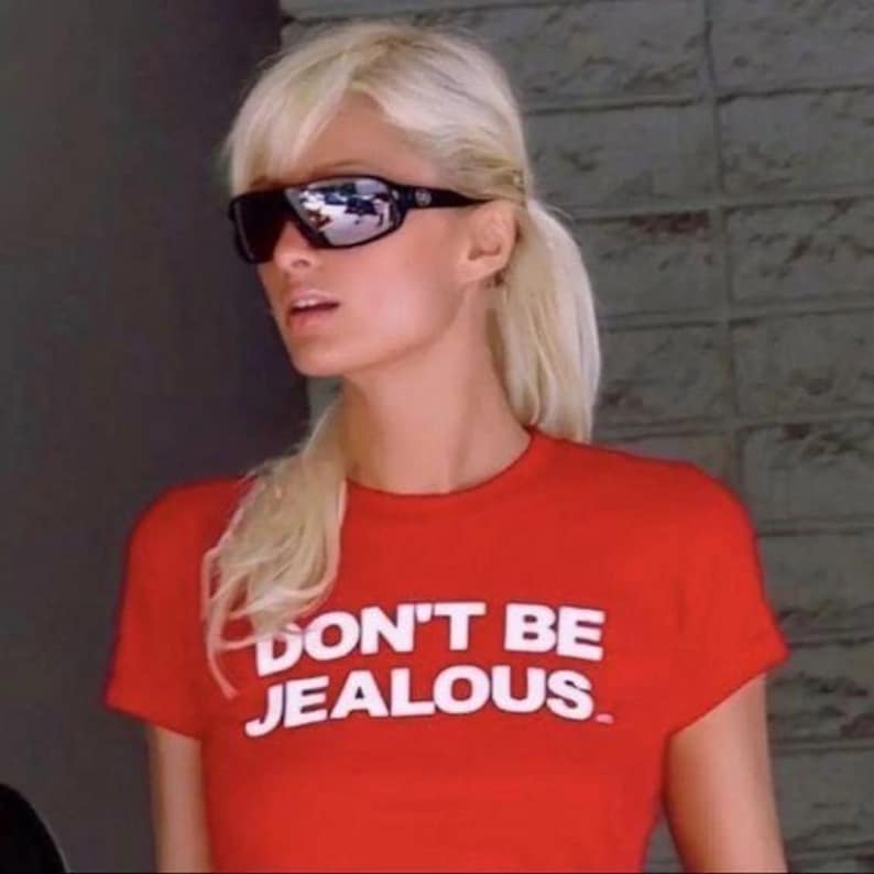 K&ouml;nnte beinhalten: Ein rotes T-Shirt mit dem wei&szlig;en Text "DON'T BE JEALOUS". Die Person auf dem Bild tr&auml;gt eine schwarze Sonnenbrille und hat blonde Haare, die zu einem Pferdeschwanz gebunden sind.