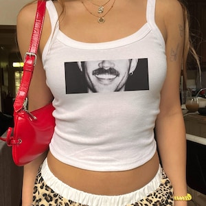May include: White vest top featuring a black and white image of a smiling face with a moustache. A red shoulder bag is on the left. The person is wearing a gold necklace and leopard print trousers.