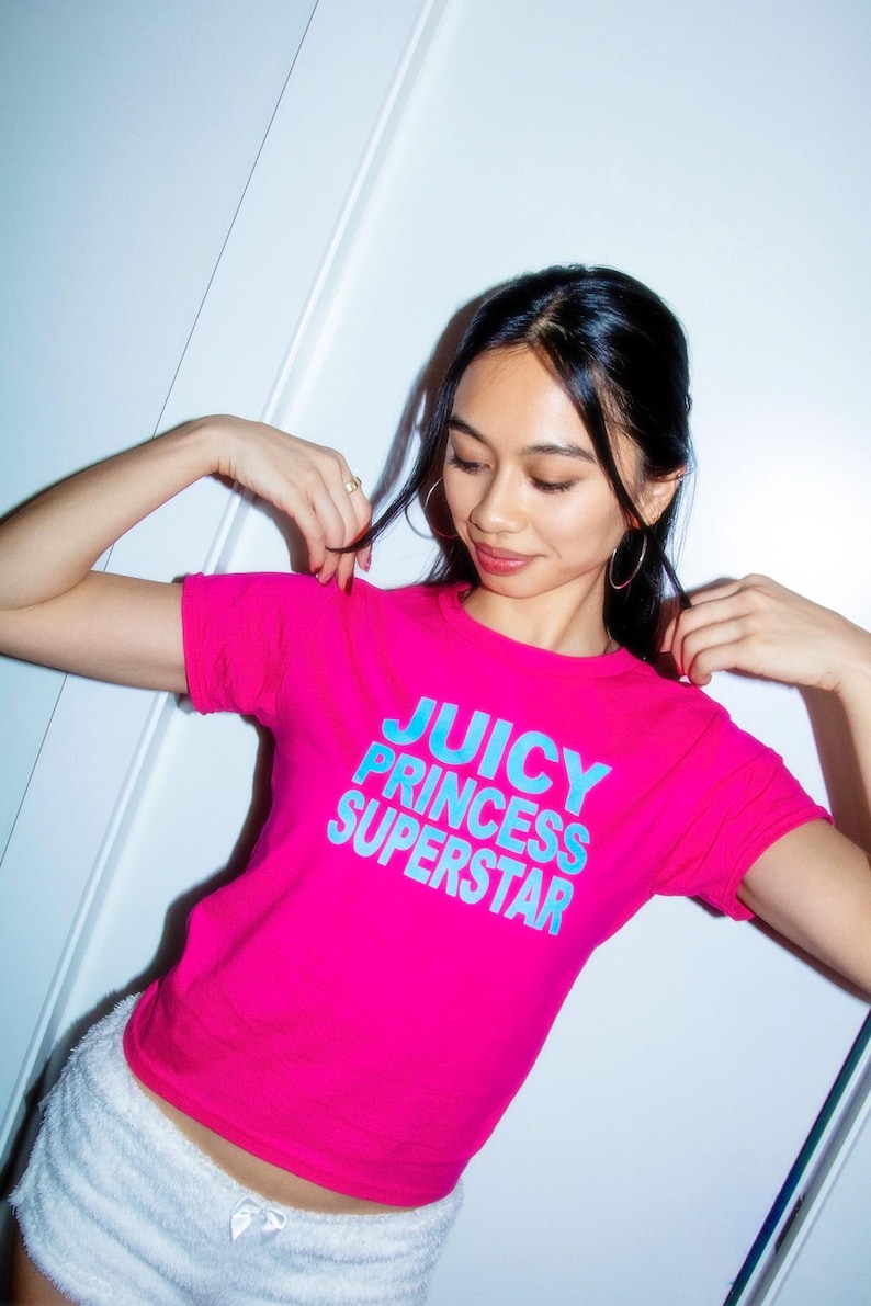 May include: A bright pink t-shirt with the text "JUICY PRINCESS SUPERSTAR" in blue. The model is wearing white shorts. The shirt is a crew neck style.