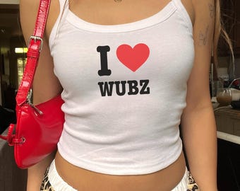Może przedstawiać: Biały top na ramiączkach z napisem "I ❤️ WUBZ" w kolorze czarnym i czerwonym. Top jest połączony ze spodniami w panterkę i czerwoną torebką na ramię. Złoty naszyjnik i bransoletka dopełniają wygląd.