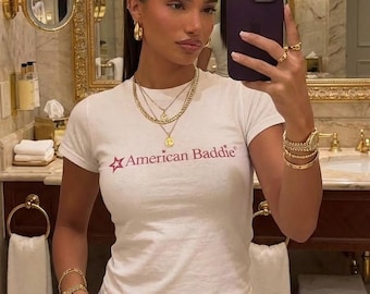 Op de afbeelding: Een wit t-shirt met de tekst "American Baddie" in roze, gecombineerd met gouden sieraden, waaronder kettingen, armbanden en ringen. De persoon houdt een paarse telefoon vast. De achtergrond toont een badkamer met gouden accenten.
