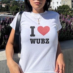 May include: White t-shirt with the text "I ❤️ WUBZ" in black and red. The shirt is short-sleeved and has a crew neck. The person in the image is wearing sunglasses and a black shoulder bag.