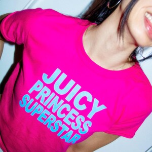 May include: A bright pink t-shirt with the words "JUICY PRINCESS SUPERSTAR" in light blue block letters. The shirt has a round neckline and short sleeves. The image is a close-up of the shirt.