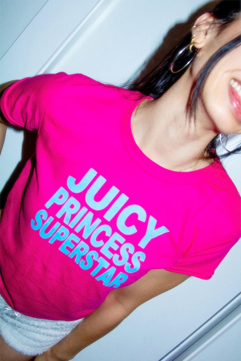 May include: A bright pink t-shirt with the words "JUICY PRINCESS SUPERSTAR" in light blue block letters. The shirt has a round neckline and short sleeves. The image is a close-up of the shirt.