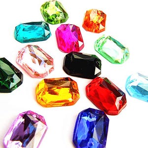May include: A selection of multi-coloured, faceted gemstone beads. The beads are in various colours, including green, blue, pink, and orange. Each bead has a rectangular shape with multiple facets, creating a sparkling effect. The beads are scattered on a white background.