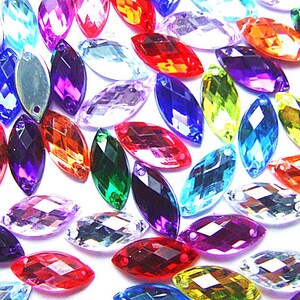 May include: A selection of multi-coloured, faceted, oval-shaped rhinestones. The rhinestones are in shades of red, blue, purple, green, and clear. Each has a hole for threading. The rhinestones are scattered on a white background.