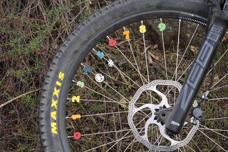 May include: Close-up of a black mountain bike wheel with a silver disc brake and a black tire with the word "MAXXIS" printed on the side. The spokes are numbered with colorful stickers.