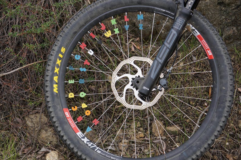 May include: A black mountain bike wheel with a disc brake and a black tyre with yellow lettering that reads "Maxxis EX471 DT Swiss". The spokes are decorated with colourful letters.