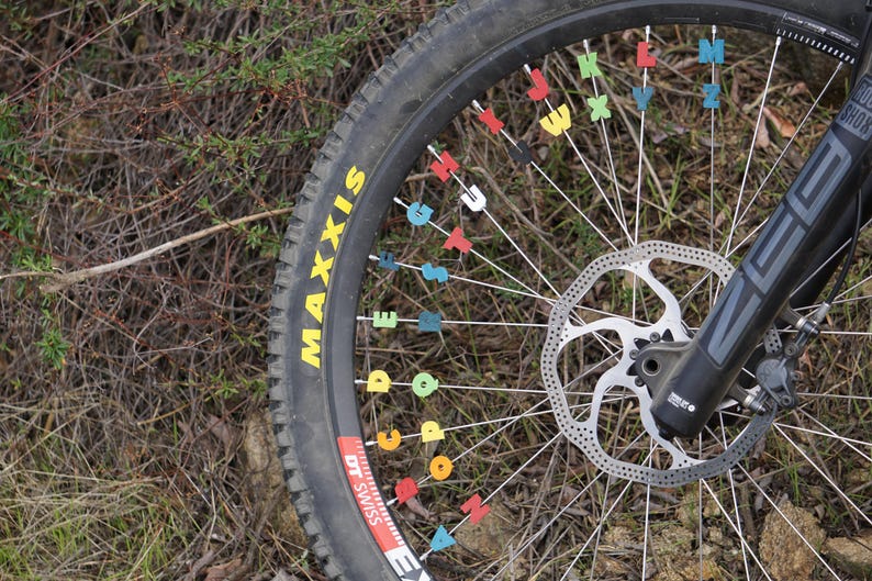 May include: A close-up of a black mountain bike wheel with a disc brake and a Maxxis brand tyre. The spokes are decorated with colourful letter stickers.
