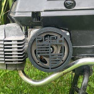 Puede incluir: Primer plano de un componente de motor negro y plateado con la palabra "PUCH" visible. El motor es parcialmente visible, con un tubo de escape plateado y una cubierta negra. El fondo es hierba verde.