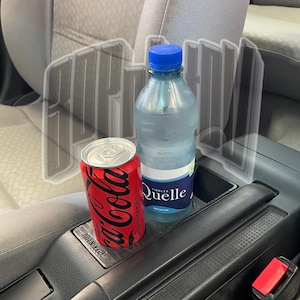 Könnte beinhalten: Eine rote Coca-Cola-Dose und eine transparente Plastikwasserflasche mit blauem Deckel und Etikett, die in der Konsole eines Autos liegen. Die Dose ist mit "Coca-Cola Zero Sugar" beschriftet. Die Wasserflasche ist mit "Quelle" beschriftet.