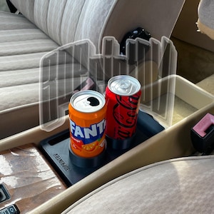Puede incluir: Interior de coche con consola central con acabado de madera y una bandeja negra con dos latas de refresco. Una lata es naranja con texto "FANTA", la otra es roja con un logotipo blanco. Asientos beige rodean la consola.