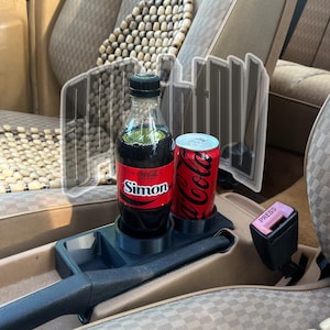 以下が含まれることがあります： 車のカップホルダーに、コカ・コーラ・シモンとコカ・コーラの缶が入っています。ボトルには白い文字の赤いラベルがあり、缶は白い文字の赤色です。車の内装はタン色で、木製のビーズのシートカバーが付いています。