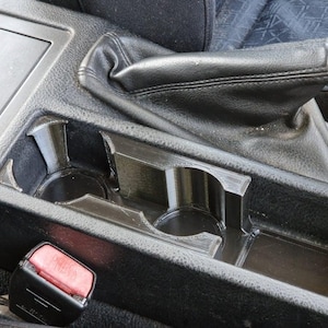 Könnte beinhalten: Nahaufnahme der Mittelkonsole eines schwarzen Autos mit zwei Getränkehaltern und einem Ablagefach. Die Konsole ist aus strukturiertem Kunststoff und hat einen schwarzen Lederschalthebelbalg.