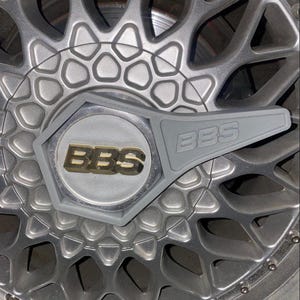 Puede incluir: Una rueda de metal gris con una tapa central hexagonal que presenta el logotipo de BBS en letras doradas. Una llave de tuercas de rueda de plástico gris con el logotipo de BBS está unida a la tapa central.