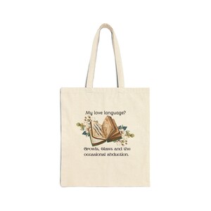May include: Cream-colored tote bag with a whimsical design. The design features an open book with floral accents and the text "My love language? Growls, Claws and the occasional abduction."