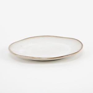 May include: A white, organically shaped ceramic plate with a slightly raised rim. The rim has a brown, textured edge. The plate is set against a white background, highlighting its simple, modern design. Ideal for serving appetizers or small dishes.