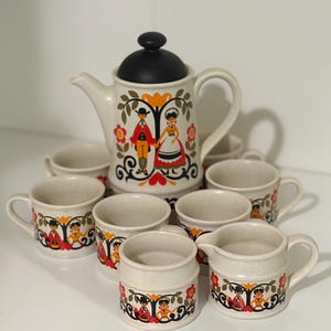 May include: A vintage tea set with a cream-colored base and colorful folk art designs. The set includes a teapot, creamer, and six cups.