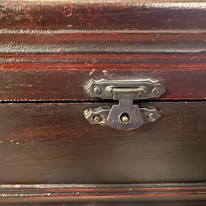 May include: Close-up of a dark brown wooden box with a metal clasp. The box has a distressed finish with visible wood grain and red accents. The metal clasp is ornate and adds a vintage feel.
