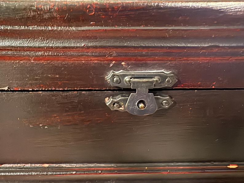 May include: Close-up of a dark brown wooden box with a metal clasp. The box has a distressed finish with visible wood grain and red accents. The metal clasp is ornate and adds a vintage feel.