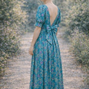 May include: A teal floral print maxi dress with a low back and a large bow detail. The dress features short sleeves and a full skirt. The floral pattern includes purple and white flowers. The dress is suitable for a special occasion.