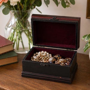 May include: A black wooden jewellery box with a burgundy velvet interior, open to reveal necklaces, bracelets, and rings. The box sits on a wooden surface. A vase of flowers and books are in the background. The box has a dark finish.