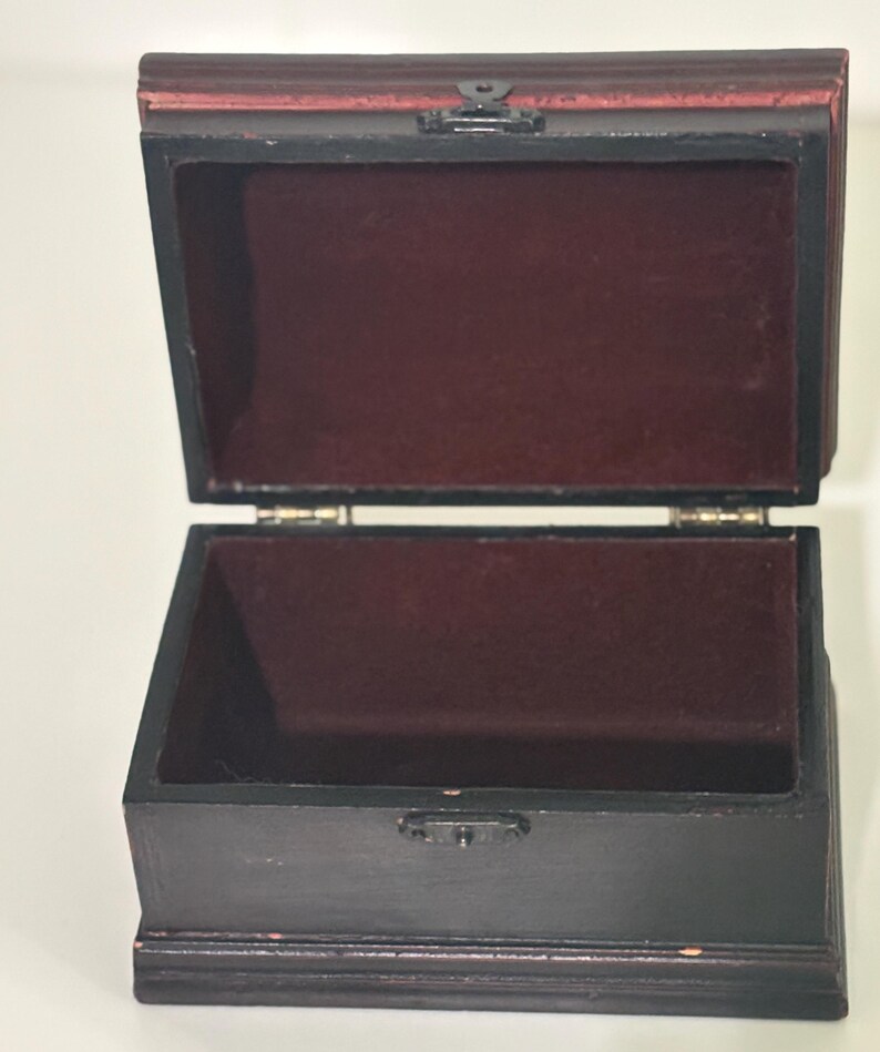 May include: A dark brown wooden box with a hinged lid, open to reveal a burgundy velvet interior. The top of the box is reddish-brown. A small metal clasp secures the lid. The box is designed for storing small items.