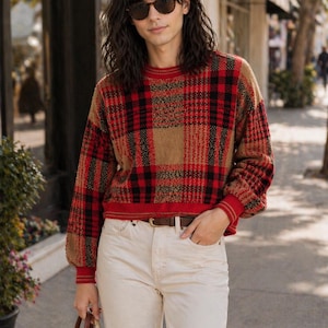 May include: A person wearing a red, black, and tan plaid sweater with a brown belt and cream trousers. The person is also wearing sunglasses and carrying a brown handbag. The sweater has a crew neck and long sleeves.