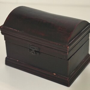 May include: A small, dark brown wooden chest with a curved lid and a rectangular base. The chest has a metal clasp and is likely used for storing small items. The wood grain is visible, and the finish has a slightly distressed look.
