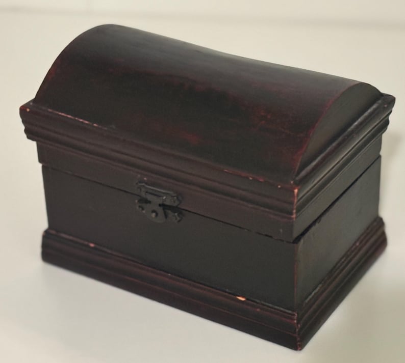 May include: A small, dark brown wooden chest with a curved lid and a rectangular base. The chest has a metal clasp and is likely used for storing small items. The wood grain is visible, and the finish has a slightly distressed look.