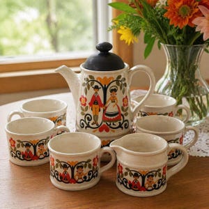Pode incluir: Um conjunto de chá de cerâmica vintage com uma chaleira com tampa preta, um cremeira e várias xícaras combinando. O conjunto é decorado com um design de arte popular de um casal em trajes tradicionais, flores e volutas decorativas.