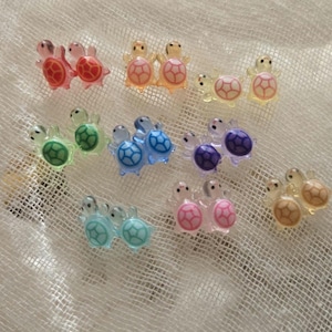 May include: A set of 10 pairs of turtle earrings in various colours, including pink, blue, green, and yellow. Each pair features two small turtles facing each other.