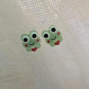 May include: Two light green frog-shaped earrings with black eyes, red hearts, and red cheeks. The earrings are small and have a cartoonish design. The background is a white, textured surface.