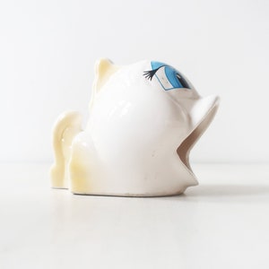 May include: White ceramic piggy bank with a blue eye and a yellow tail. The piggy bank has an open mouth.