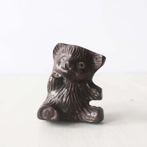 May include: A small, brown, ceramic bear figurine with a textured surface. The bear is sitting with its paws tucked in and has a cute, cartoon-like face.