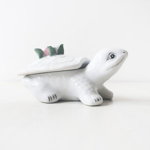 May include: A white ceramic turtle trinket box with a hinged lid. The turtle has a green and pink flower on its back.