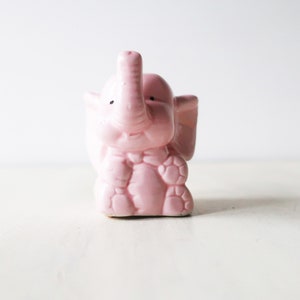 Peut inclure: Une figurine d'éléphant en céramique rose avec des yeux noirs. L'éléphant est assis, sa trompe levée et ses pattes repliées sous lui.