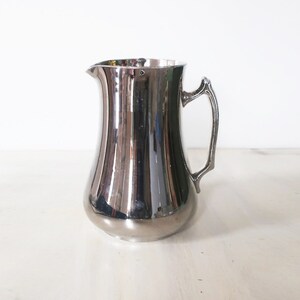 May include: A silver pitcher with a curved handle. The pitcher is made of polished metal and has a shiny, reflective surface.