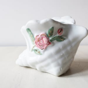 May include: White ceramic planter with a textured surface and a rose design. The pink rose has gold accents and is surrounded by green leaves. The planter has a unique, sculpted shape.