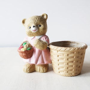 May include: A ceramic bear figurine wearing a pink dress and holding a basket of flowers. The bear is next to a small, beige, ceramic planter shaped like a woven basket.