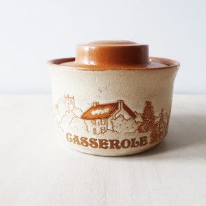 May include: A brown and white ceramic casserole dish with a lid. The dish features a cottage scene with a thatched roof and the word "CASSEROLE" printed on the front.