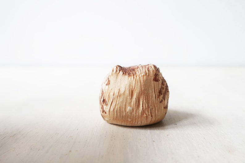 Op de afbeelding: Een klein, getextureerd, beige decoratief object met bruine accenten. Het object heeft een ruwe, organische uitstraling, mogelijk lijkend op een natuursteen of een stuk aardewerk. Het staat op een lichtgekleurd oppervlak.
