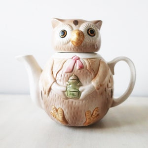 Peut inclure: Théière en céramique en forme de hibou avec un couvercle amovible. La théière est principalement beige avec des accents marron et crème. Elle présente un nœud papillon rose et tient un objet vert. Les yeux du hibou sont grands et jaunes.