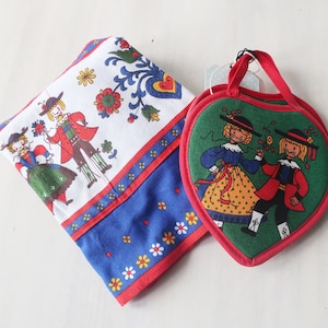 May include: Two red heart-shaped pot holders with a green background and a white design of a couple in traditional folk costumes. The pot holders have a red loop handle.