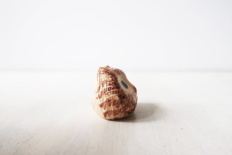 Op de afbeelding: Een kleine keramische uilfiguur in bruin- en beigetinten, met een textuur. De uil heeft een klein blauw oog en staat tegen een witte achtergrond.